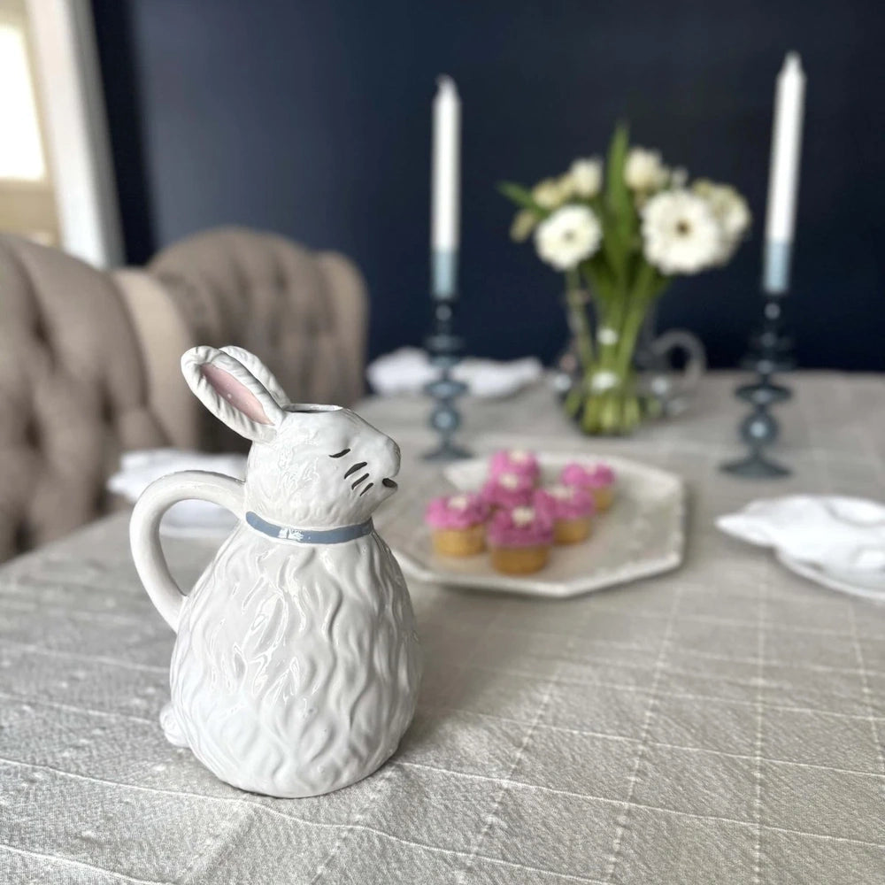 A white ceramic bunny pitcher sits on a dining table set with cupcakes, candles, and flowers.