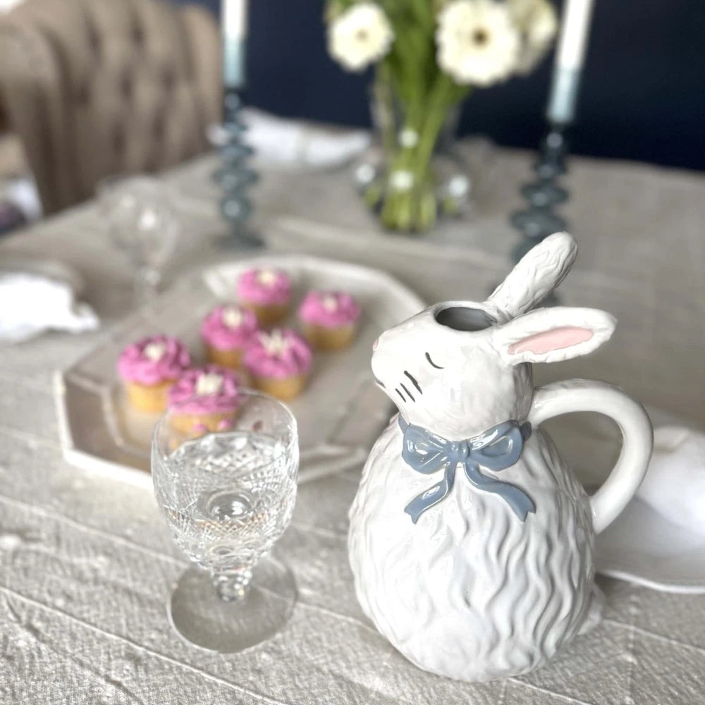 White ceramic bunny pitcher with a blue bow on a table next to a glass and pink cupcakes.