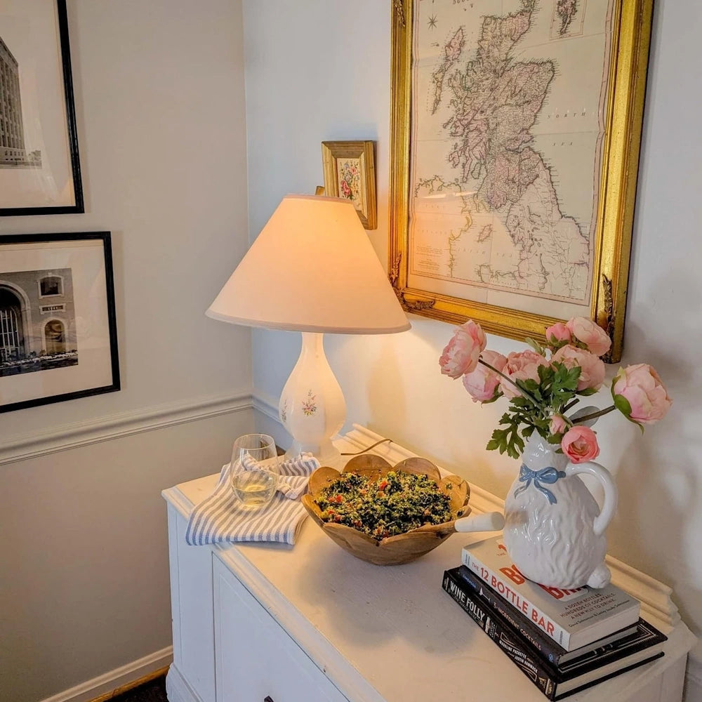 A lit table lamp, a bowl of salad, and pink flowers on a white credenza beneath framed wall art.