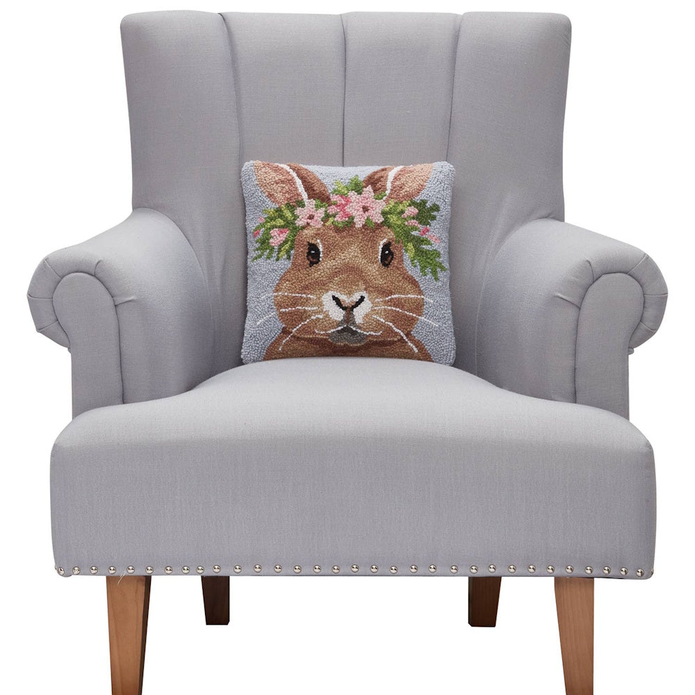 Brown bunny with pink floral crown hooked pillow on a grey tufted armchair with nailhead trim and wooden legs.