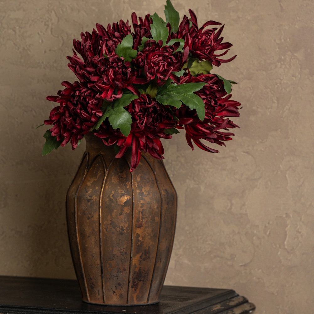 Deep burgundy mum flowers with green leaves arranged in a textured bronze vase against a neutral wall.
