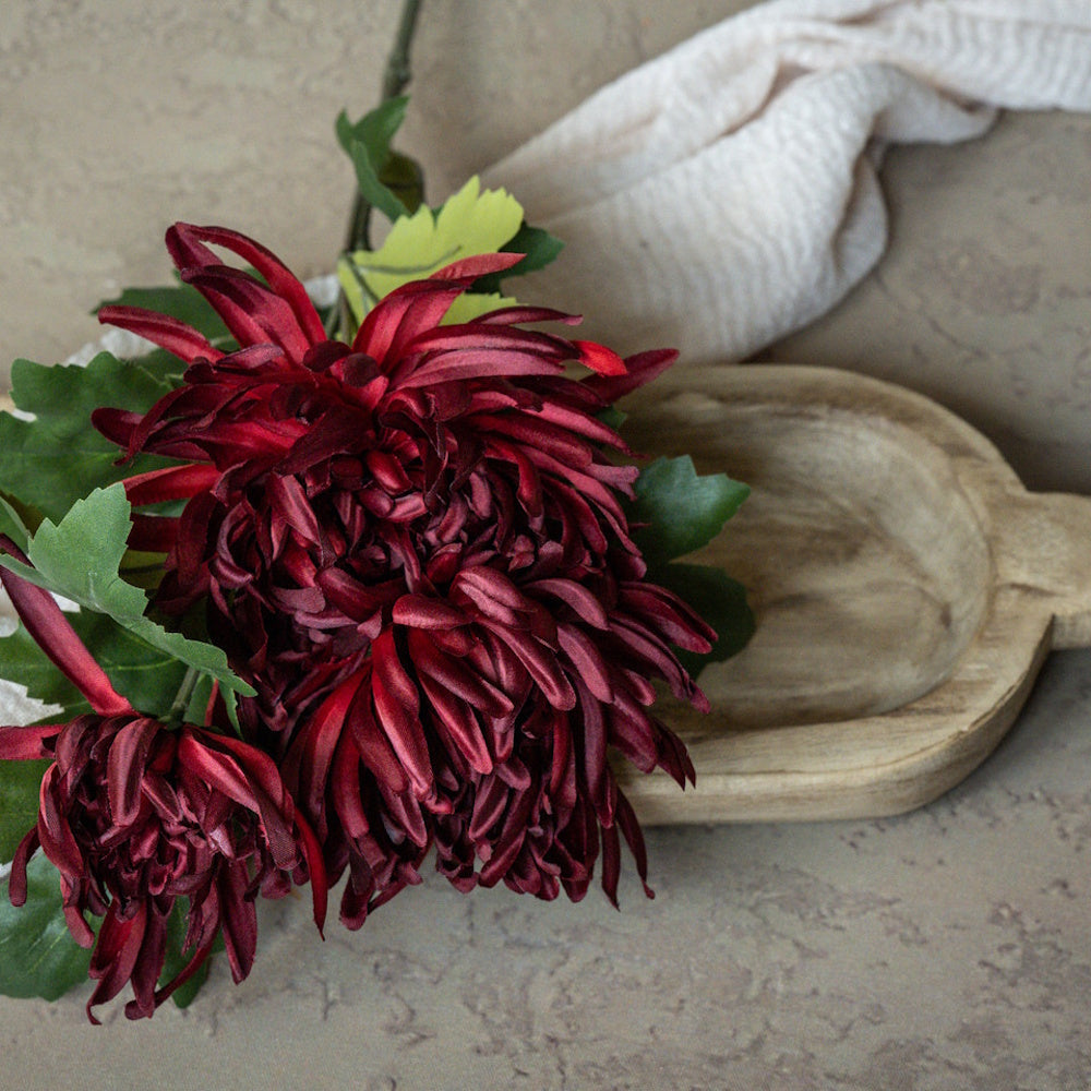 A faux burgundy chrysanthemum spray resting beside a carved wooden dough bowl and neutral cloth.