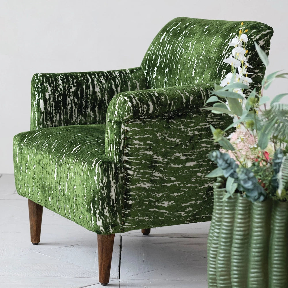 A unique armchair upholstered in green velvet with white streaking, with wooden legs and a vase of flowers beside it.