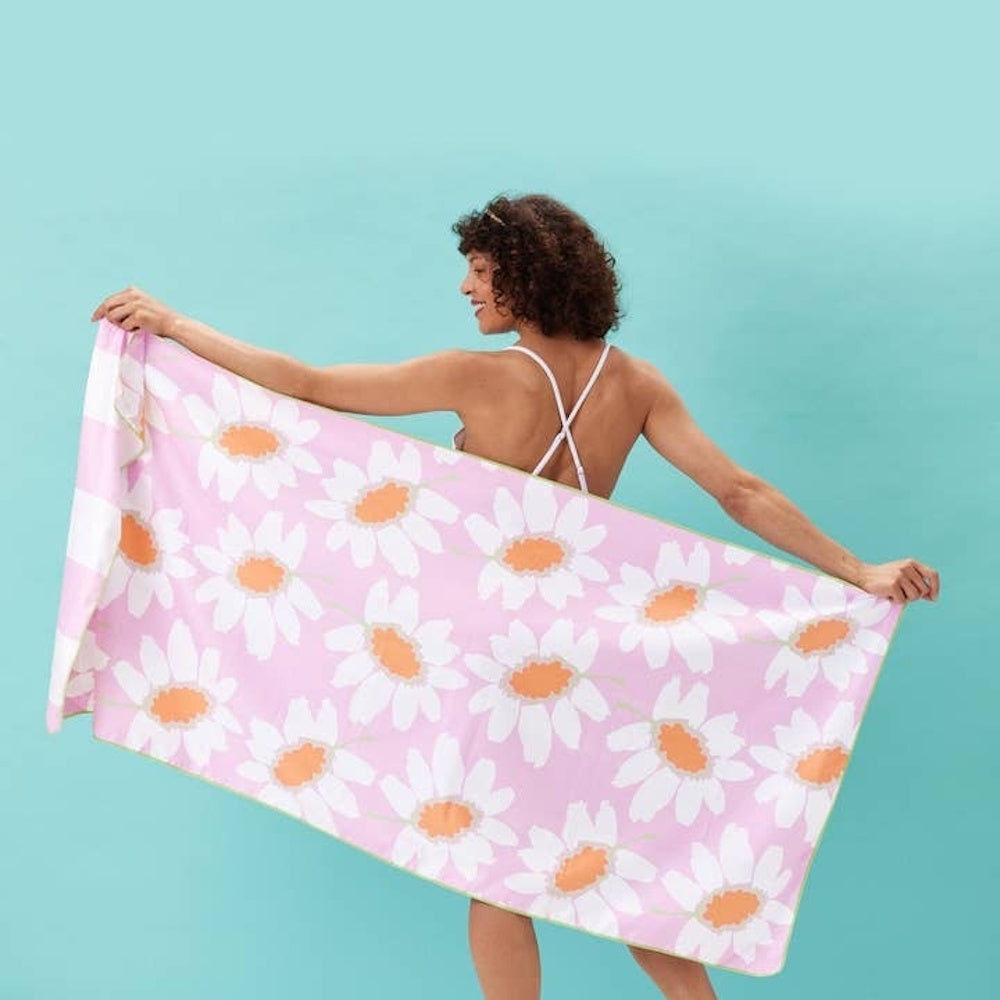 A woman smiling from behind holds up a pink beach towel patterned with large white and orange daisies.