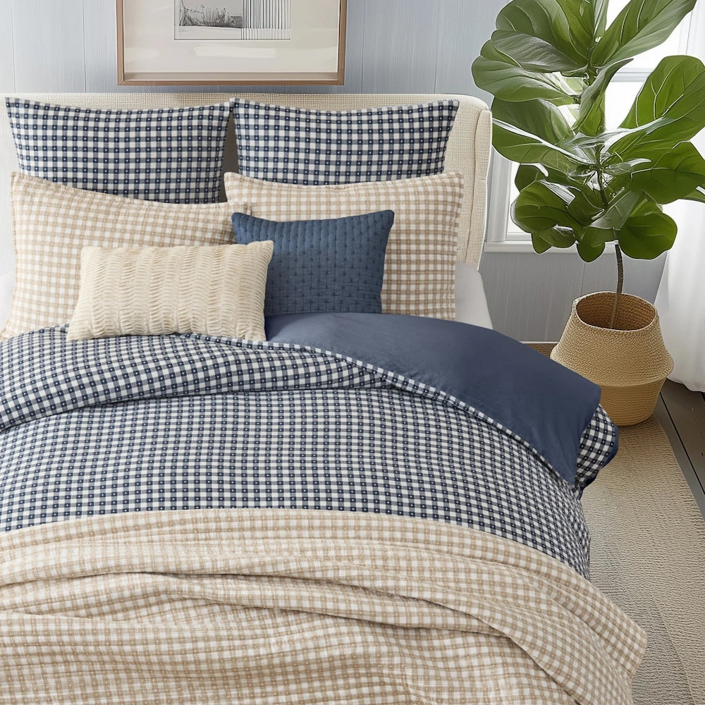 Bed styled with navy and tan gingham bedding, layered shams, accent pillows, and a potted plant.