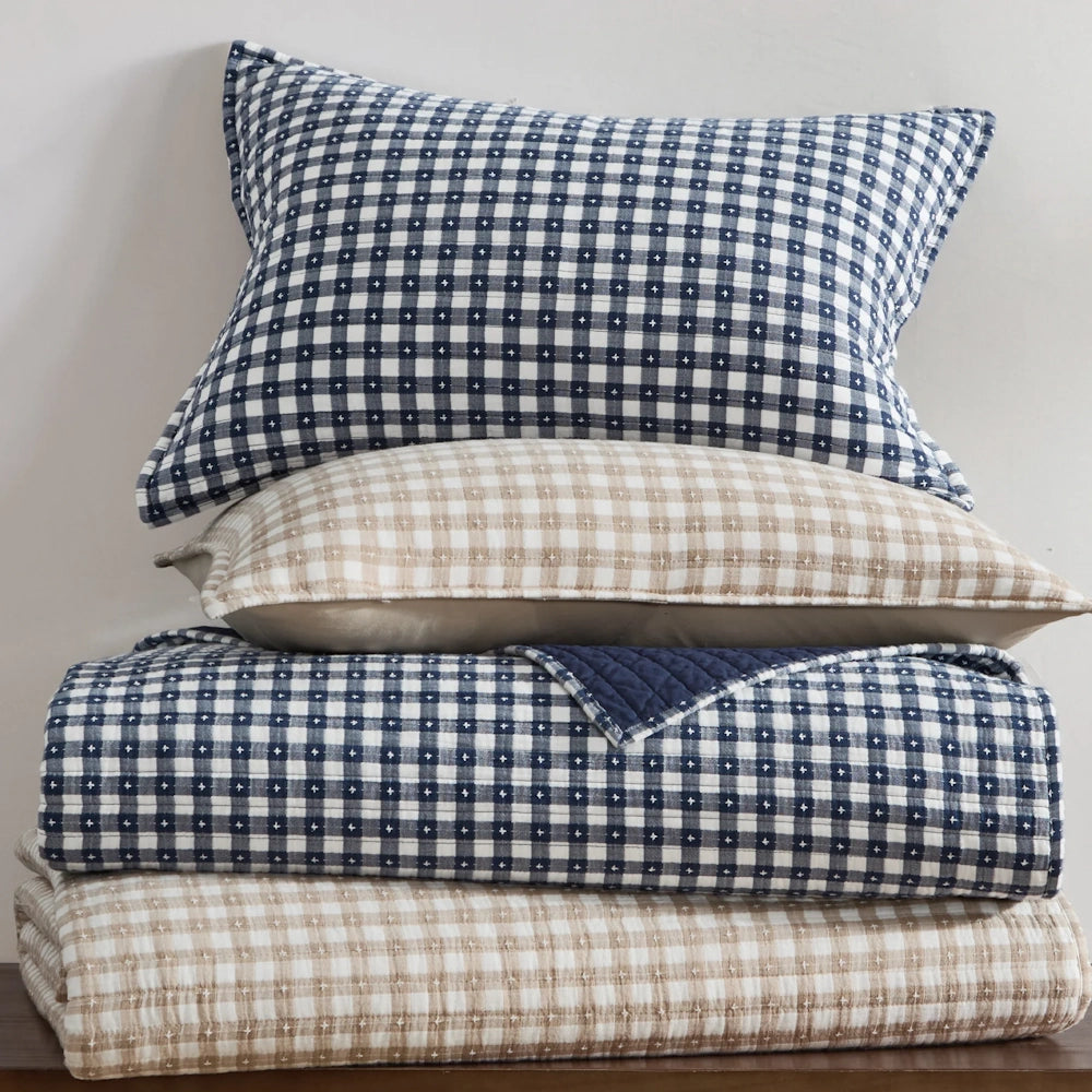 Stacked navy and beige gingham pillows and folded quilts featuring stitch detailing.