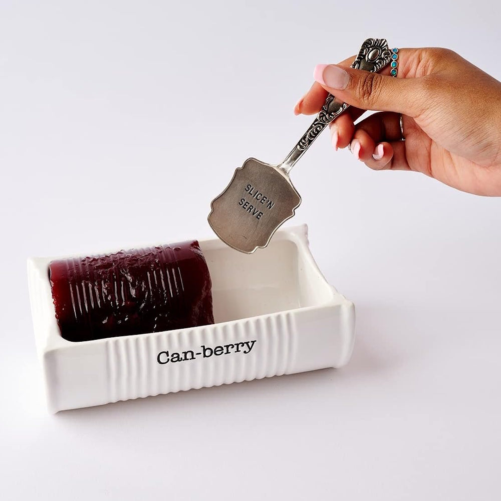 Hand holding a silver server above a white ceramic "Can-berry" dish with jellied cranberry sauce.