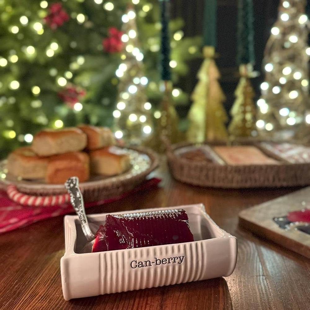 White ceramic "Can-berry" dish with cranberry sauce on a holiday table in front of Christmas tree lights.