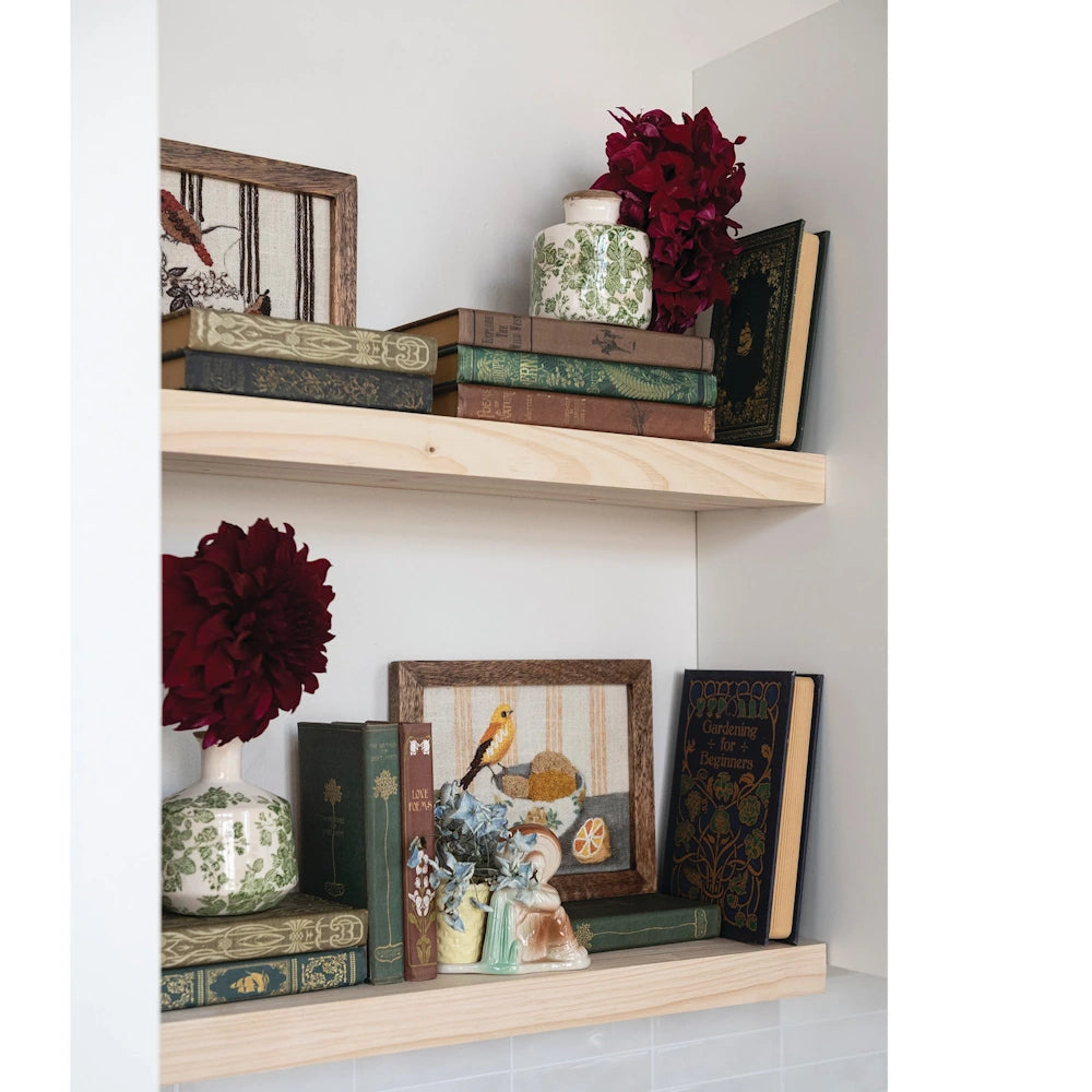 Vintage books, floral vases, and framed art arranged on light wood floating shelves with a cottagecore aesthetic.