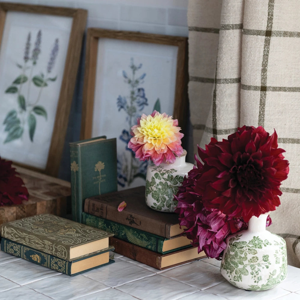 Vintage books and dahlia flowers in floral vases on a tiled surface with framed botanical prints and a linen curtain.