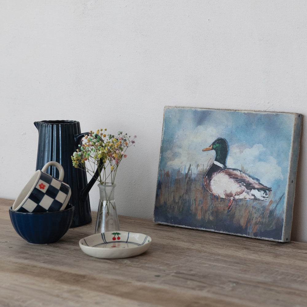Vintage mallard duck canvas painting styled on a wooden tabletop next to blue ceramics and small flowers.