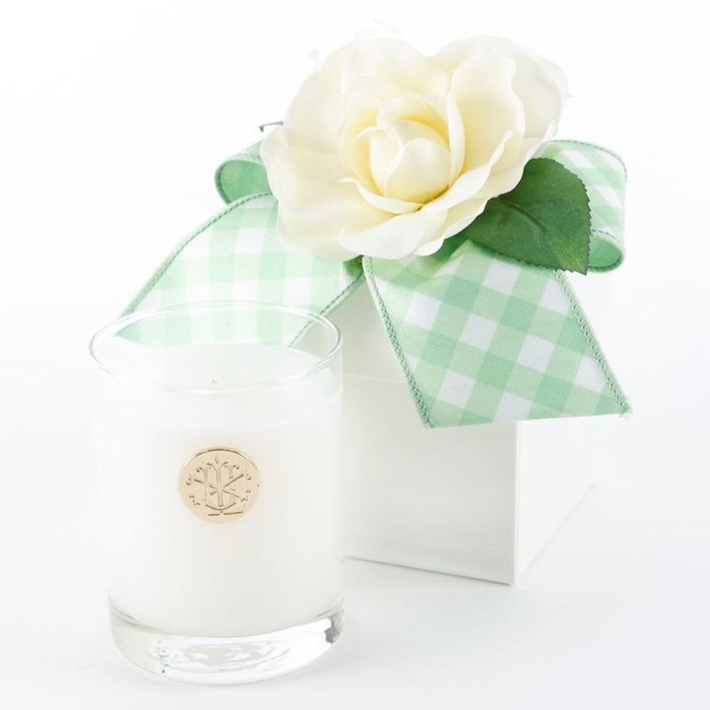White jar candle next to a white gift box topped with a green checkered bow and a white flower.
