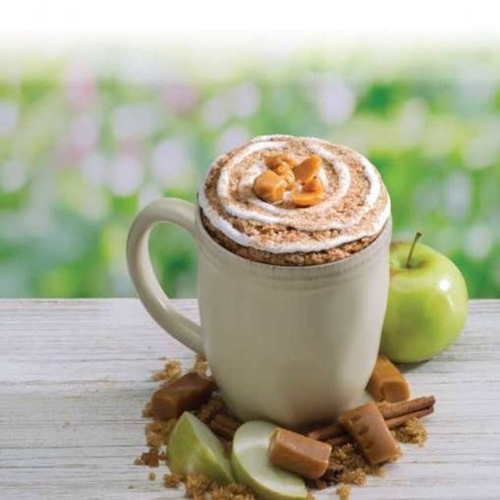 Caramel apple mug cake topped with cream, set beside green apple slices, caramel candies, and cinnamon.