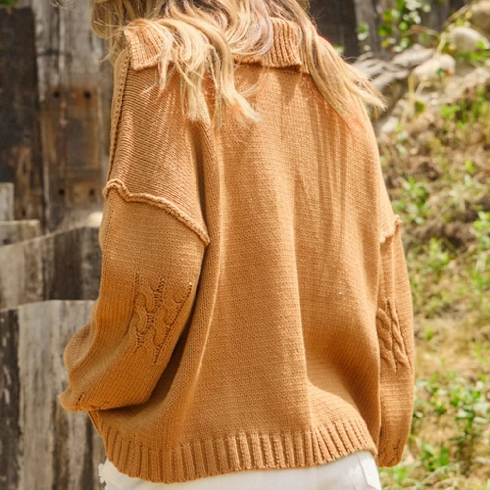 Back view of a woman wearing a caramel knit sweater with exposed seams and cable details on the sleeves.