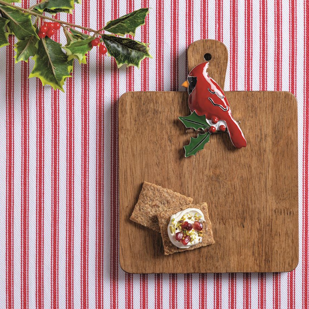 Red cardinal charm on a wooden cutting board with crackers, on a red and white striped tablecloth.