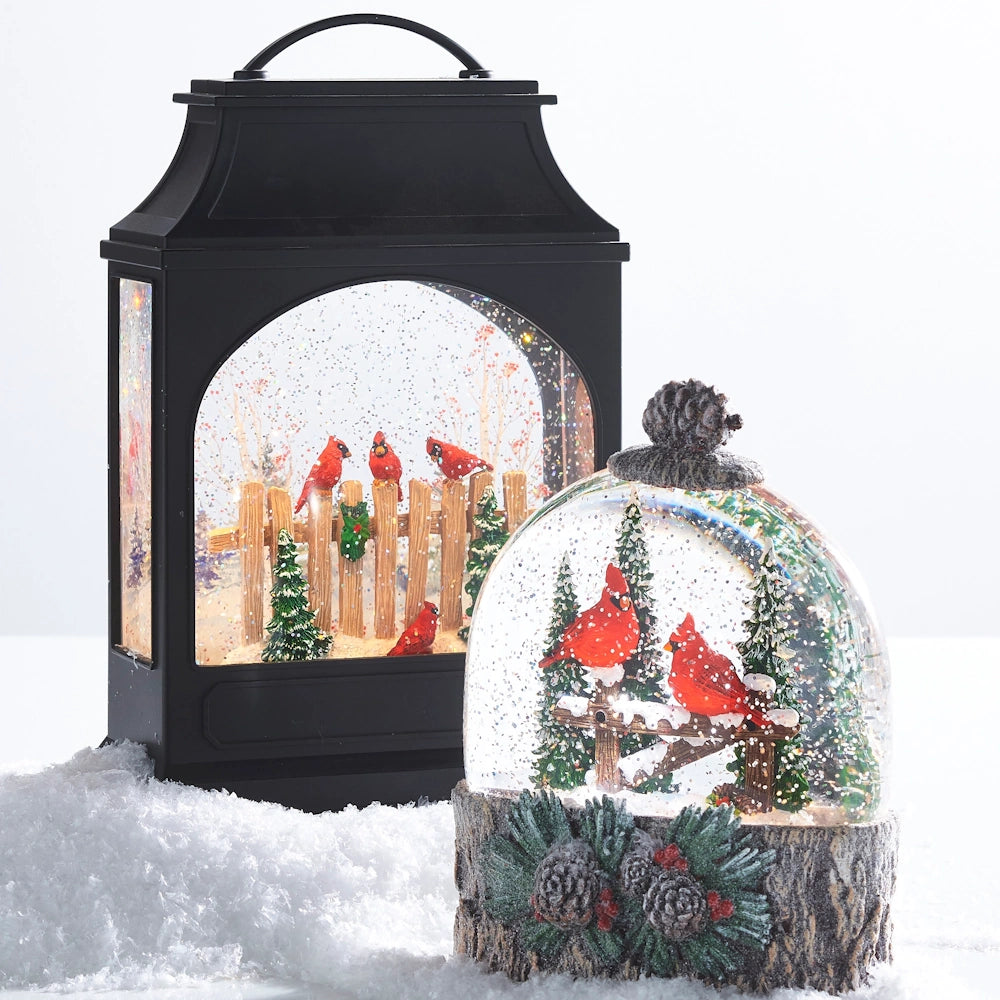 A black lighted water lantern and a dome-shaped snow globe with winter cardinals, surrounded by artificial snow.