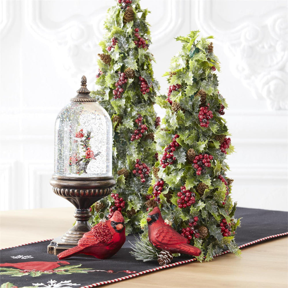 Cardinal snow globe on a pedestal next to small frosted tabletop trees and red bird ornaments.
