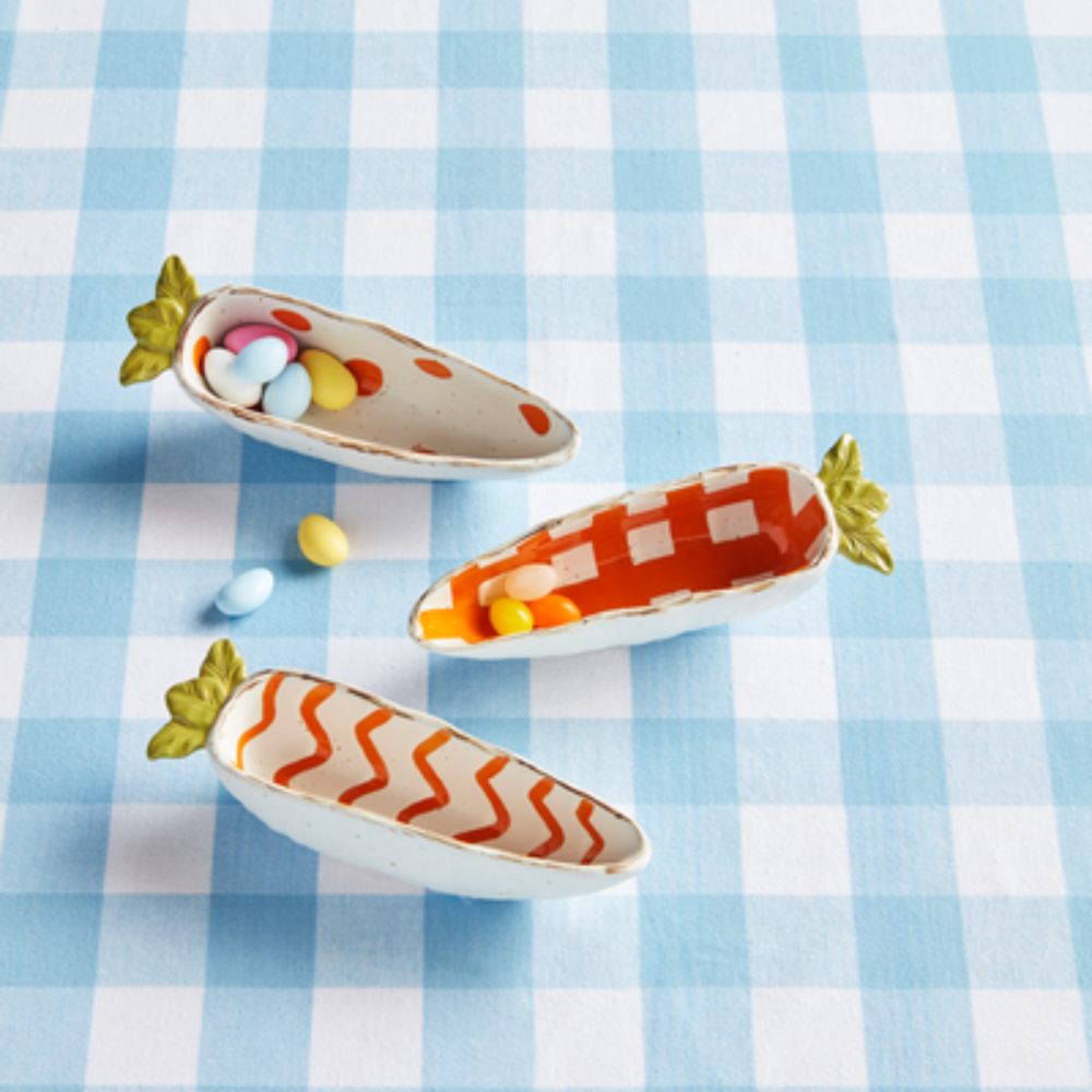 Three carrot-shaped dishes with patterned orange interiors hold pastel candies on a blue checkered tablecloth.