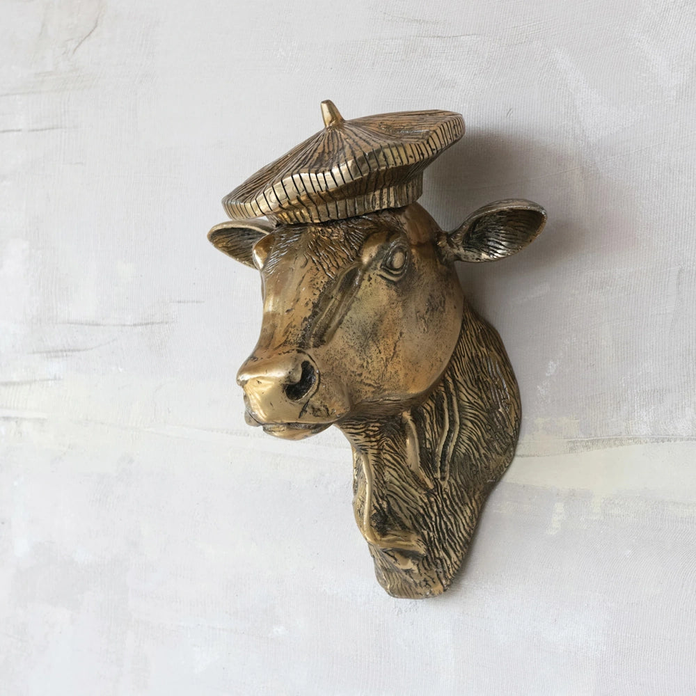 Bronze cow head wall sculpture wearing a beret mounted on a textured, light-grey wall.