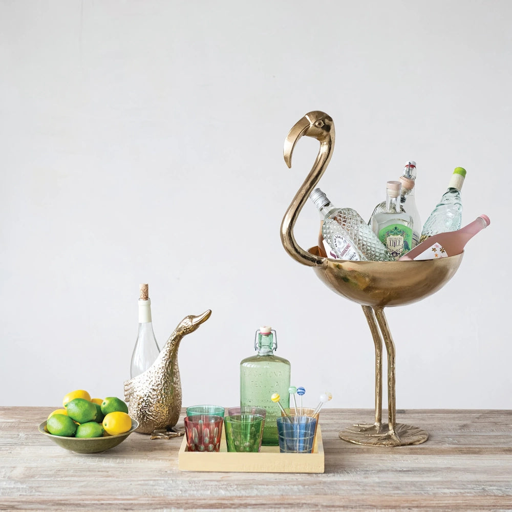 Gold duck bottle holder holding a wine bottle on a bar table next to a brass flamingo ice bucket.