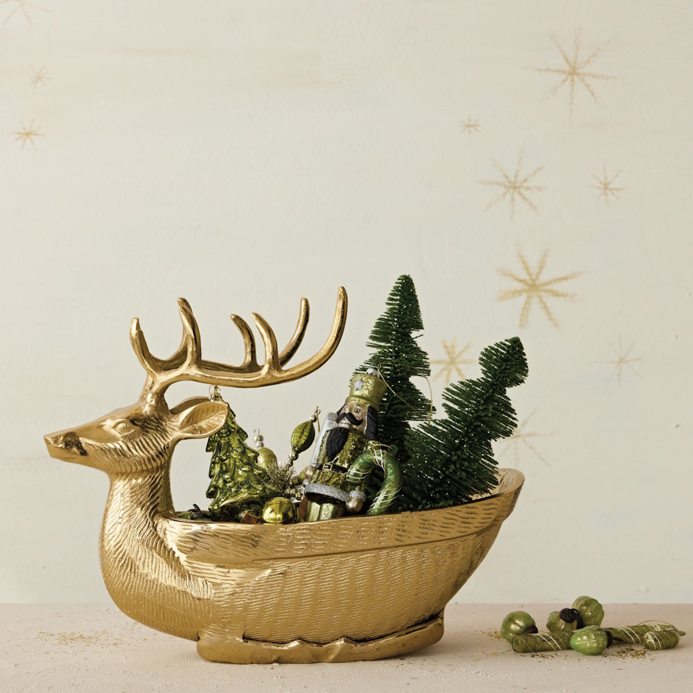 A gold reindeer-shaped bowl filled with bottle brush trees, a nutcracker, and ornaments, with star prints on the wall behind.