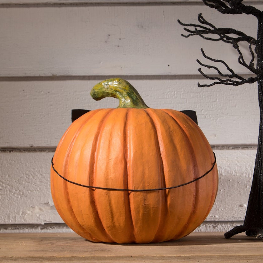 Back view of an orange pumpkin with an elastic strap holding a mask on, next to a black tree.