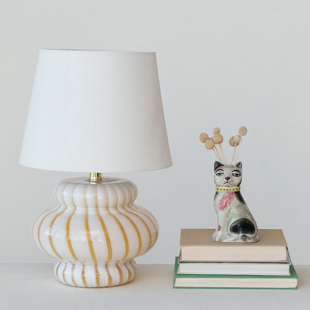 A table lamp with a striped glass base next to a small cat-shaped vase sitting on a stack of three books.