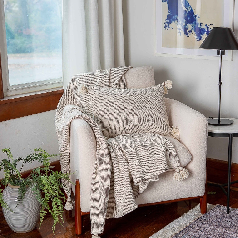 Cozy cream armchair styled with a matching patterned throw blanket, tassel pillow, lamp, and potted fern.
