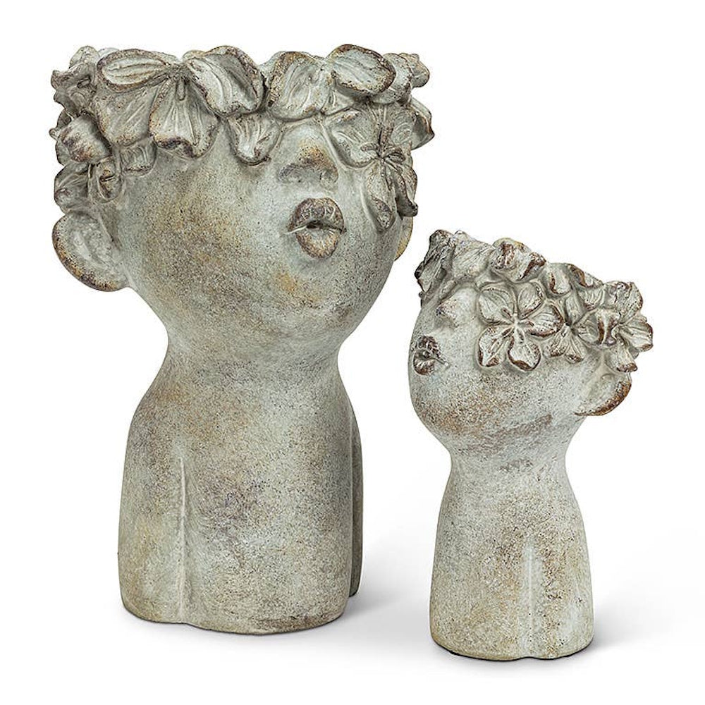 Two cement head-shaped planters with floral crowns and pucker lips in a kissing pose.