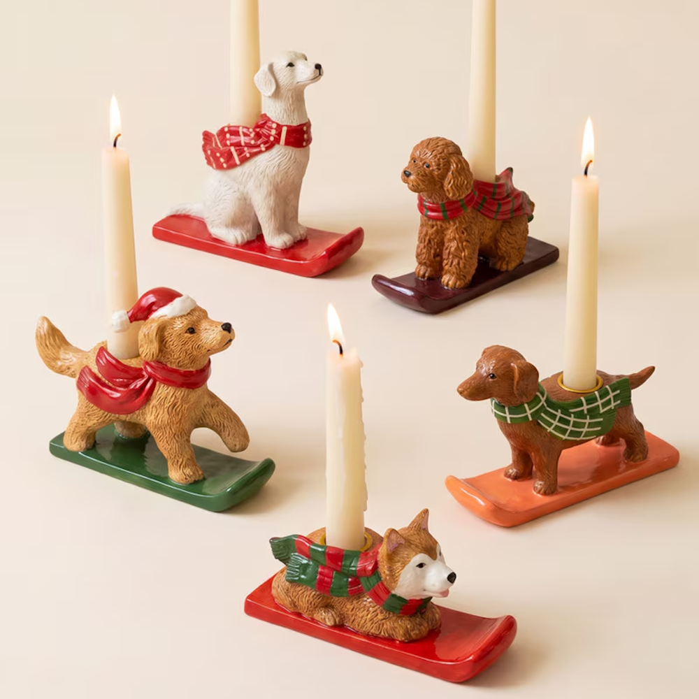 Five ceramic Christmas dog taper candleholders on sled bases with lit candles against a light background.