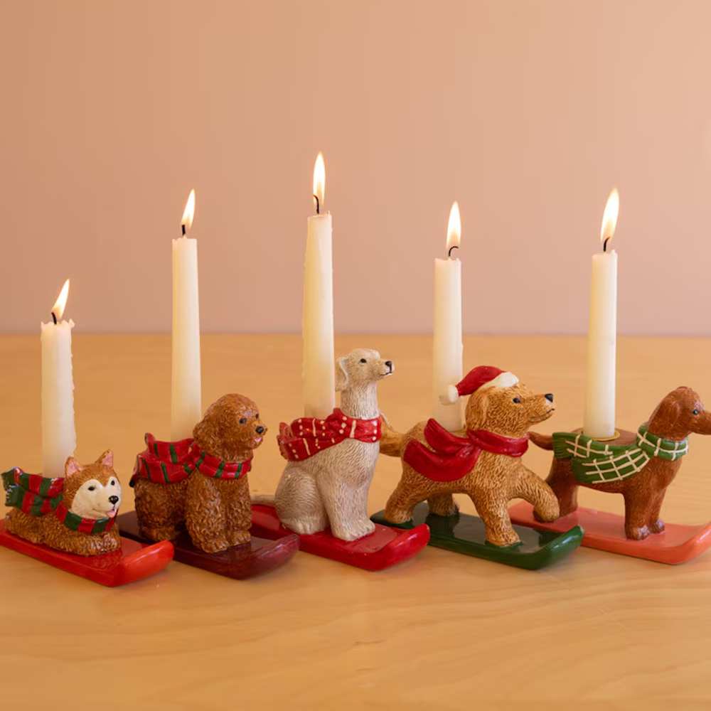 Row of five ceramic holiday dog taper candleholders on sled bases with lit white candles.