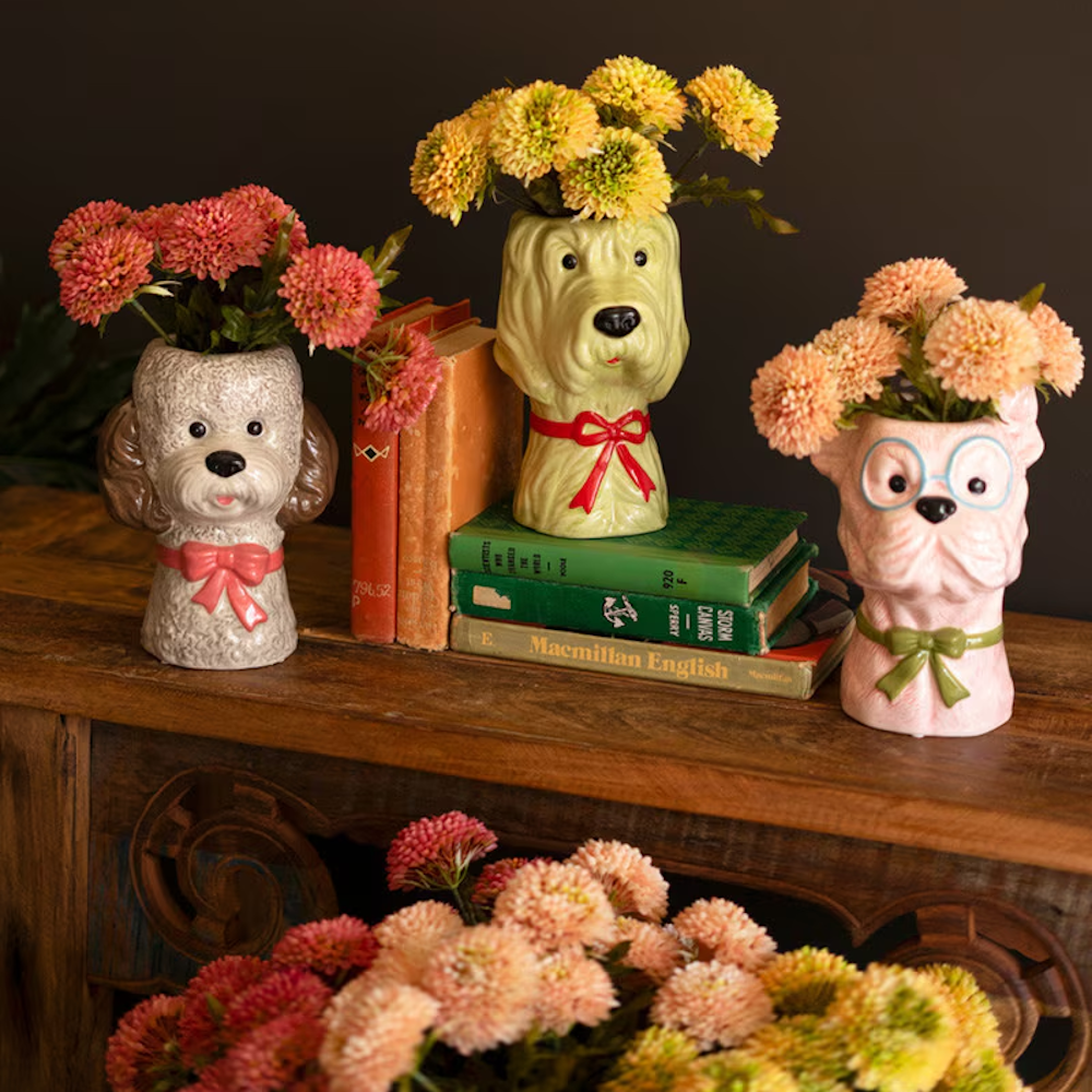 Three whimsical ceramic dog-head vases with bows and colorful flowers arranged on a wooden table with vintage books.