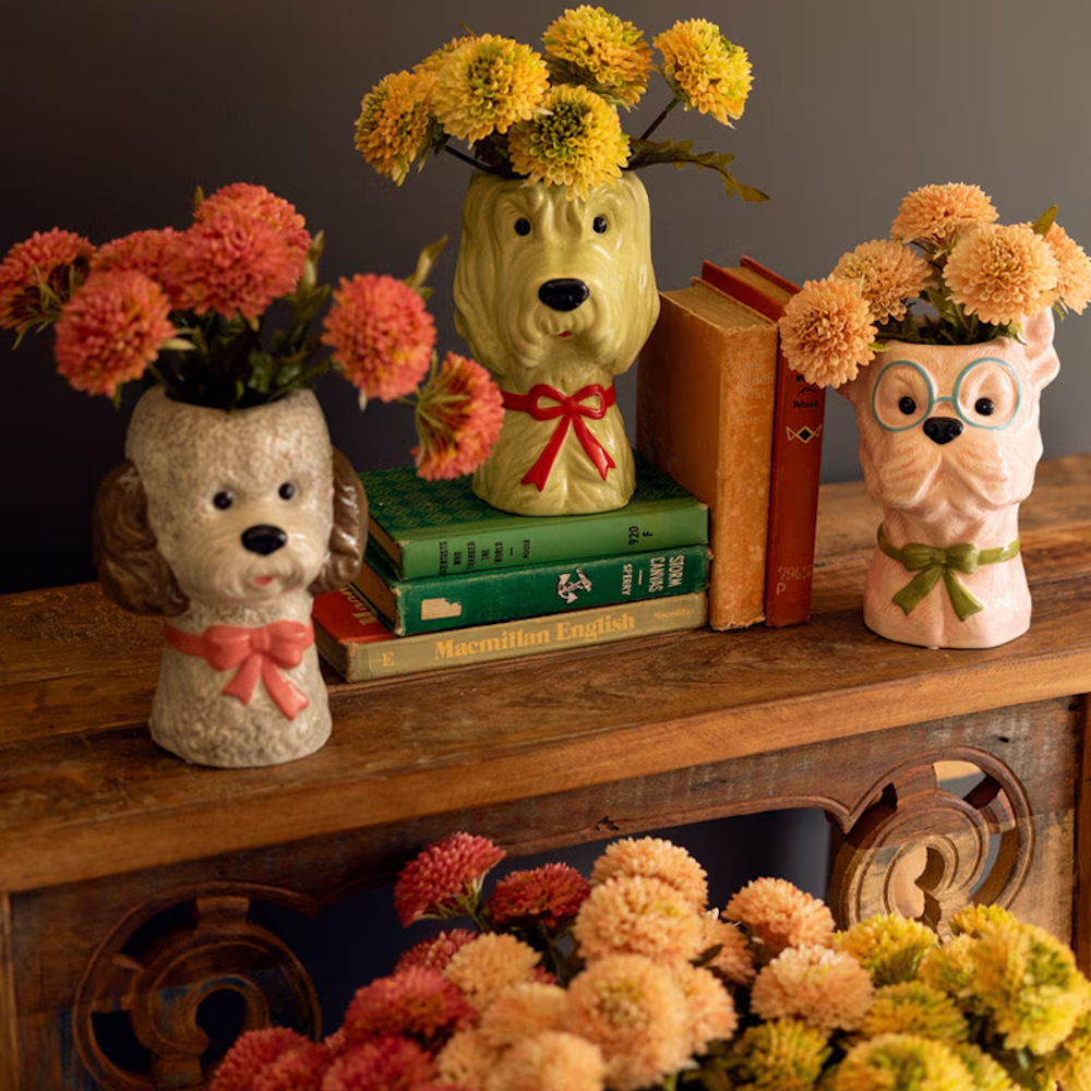Three vintage-style ceramic dog head vases with bows and flowers on a wooden table with books.
