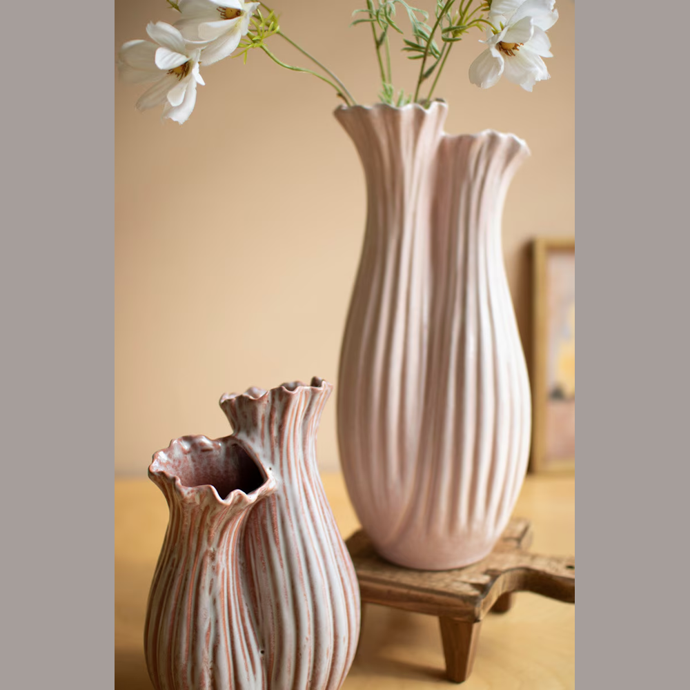 Two pink fluted ceramic vases with ruffled edges holding white flowers on a wooden stand.