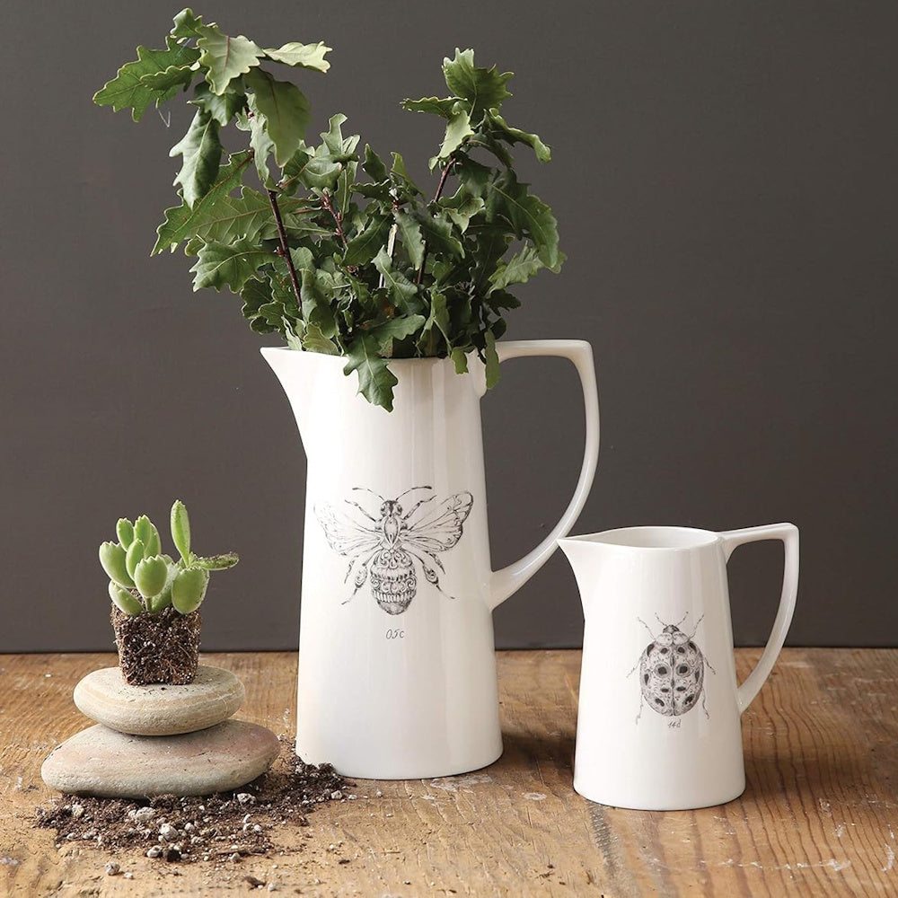 Two white ceramic pitchers with insect illustrations on a wooden table, one holding green leafy branches.