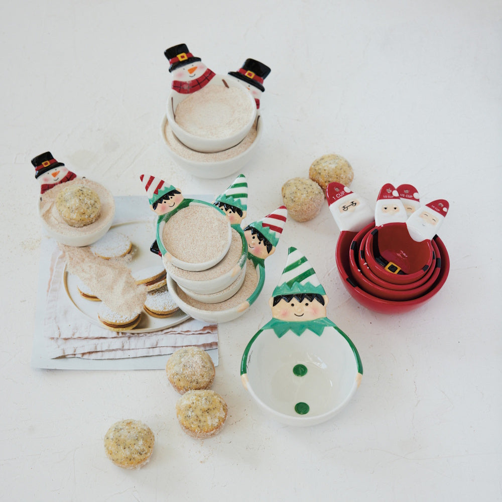 A flat lay showing various holiday-themed ceramic measuring cups shaped like Santa, elves, and snowmen, surrounded by small baked treats.