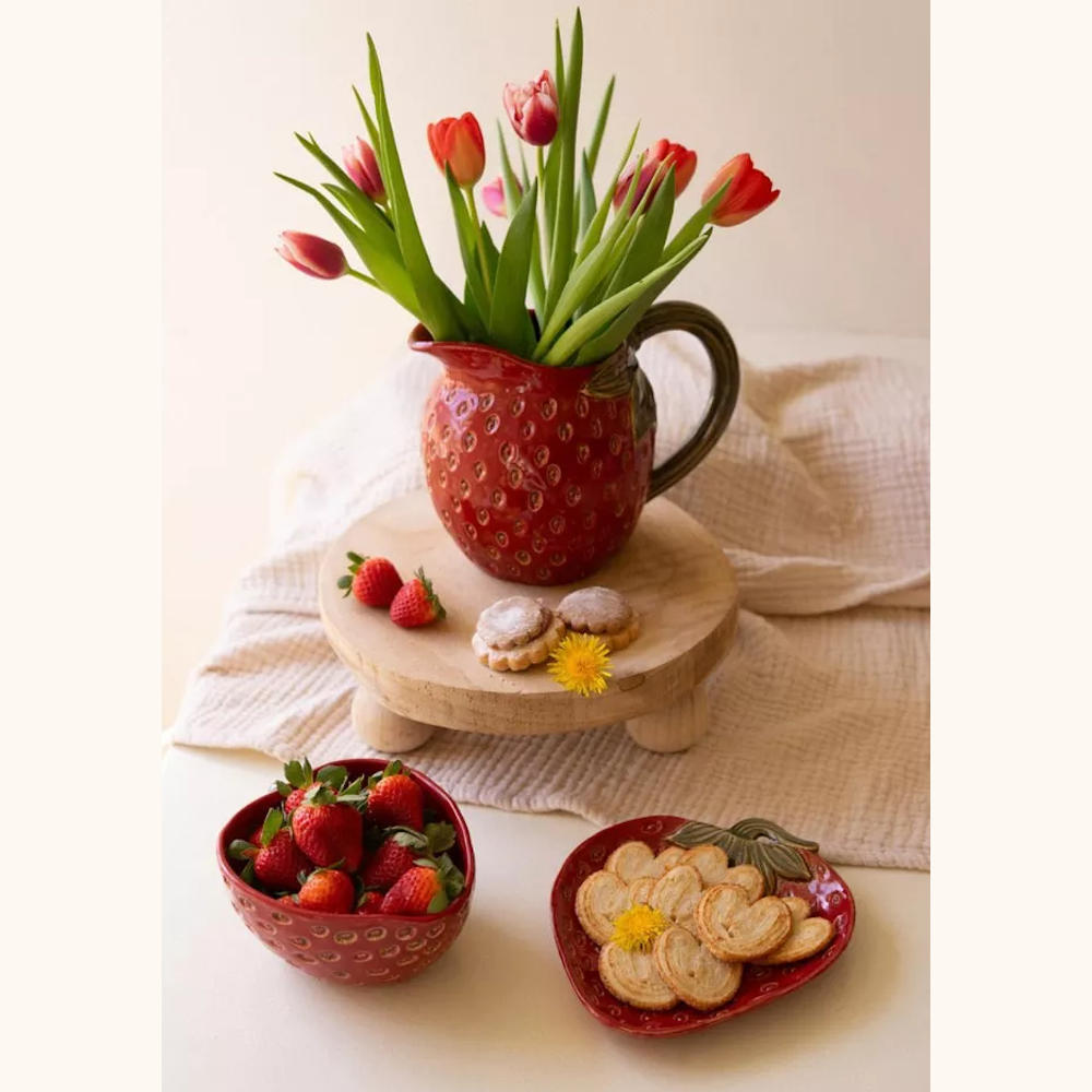 Red ceramic strawberry pitcher with tulips, bowl of strawberries, and cookies on a wooden stand and table.