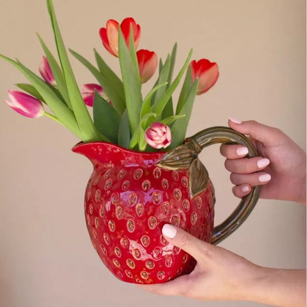 Red ceramic strawberry-shaped pitcher filled with pink and orange tulips, held by two hands against a neutral background.