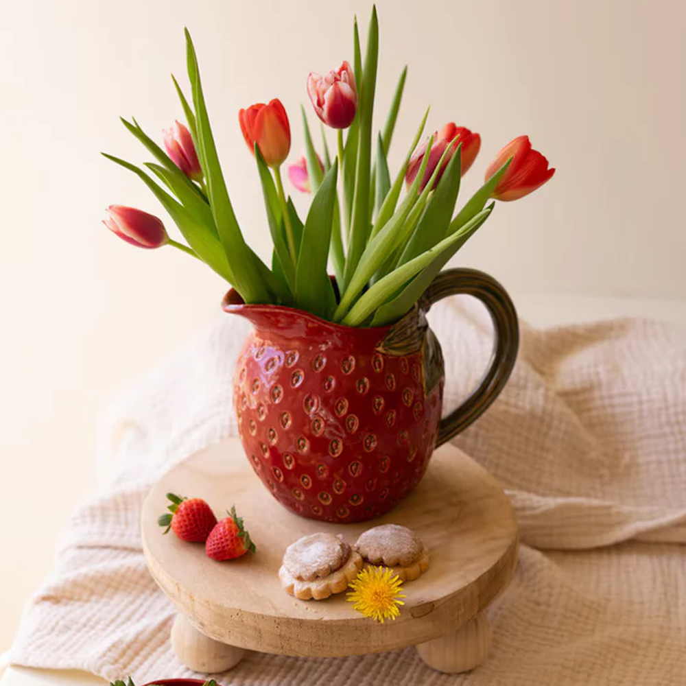 Red ceramic strawberry pitcher vase with pink tulips on a wooden riser with fresh strawberries and cookies.