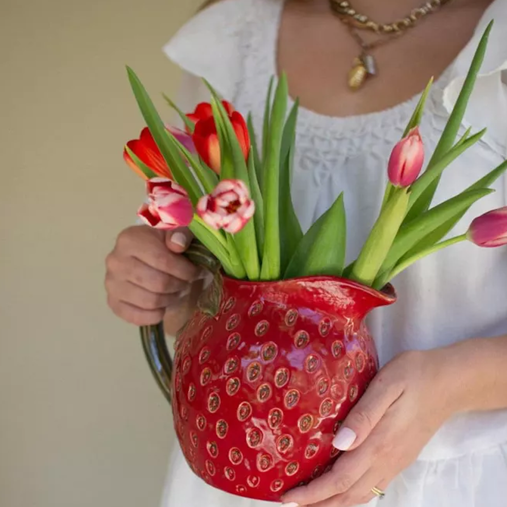 A person holding a red ceramic strawberry-shaped pitcher filled with fresh pink and red tulips.