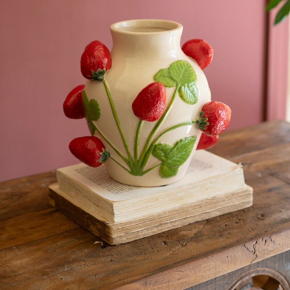 Cream ceramic vase with 3D embossed red strawberries and green leaves, sitting on vintage books on a wooden table.
