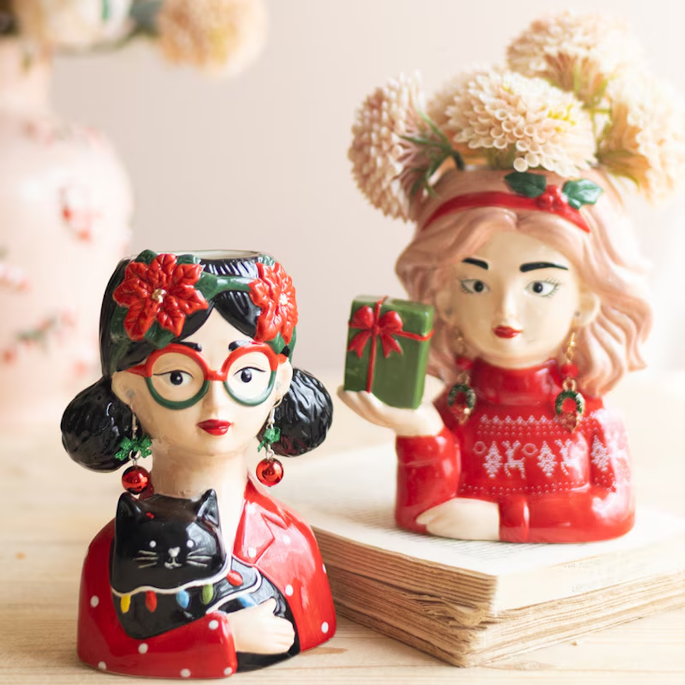 Two festive ceramic lady head vases styled on a wooden surface with flowers and a book.