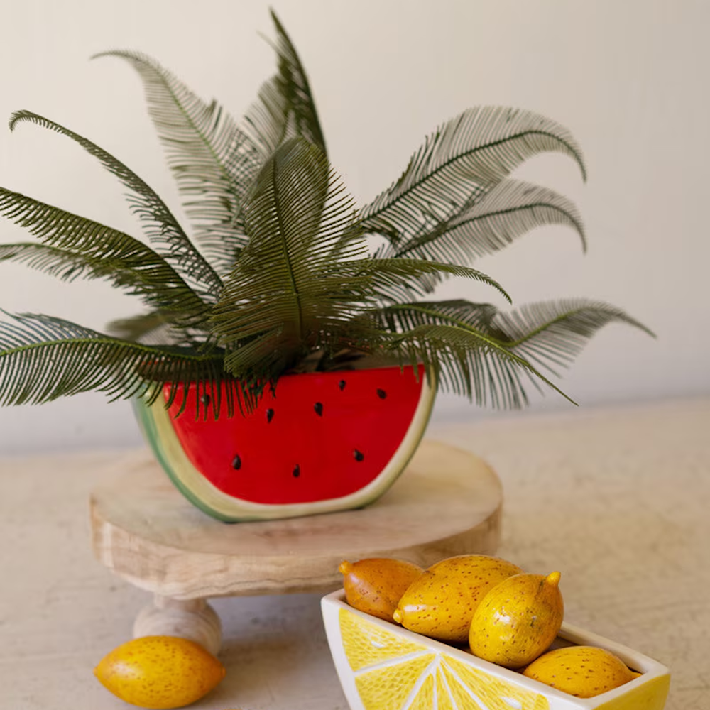 Ceramic watermelon wedge planter with a green palm-like plant, displayed on a wooden riser next to a lemon-shaped bowl.