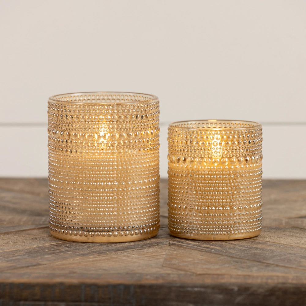 Two lit champagne-gold beaded glass candle holders of varying sizes on a wooden table.