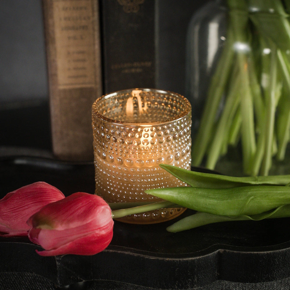 Lit candle in a champagne dotted glass vessel placed next to pink tulips and vintage books.