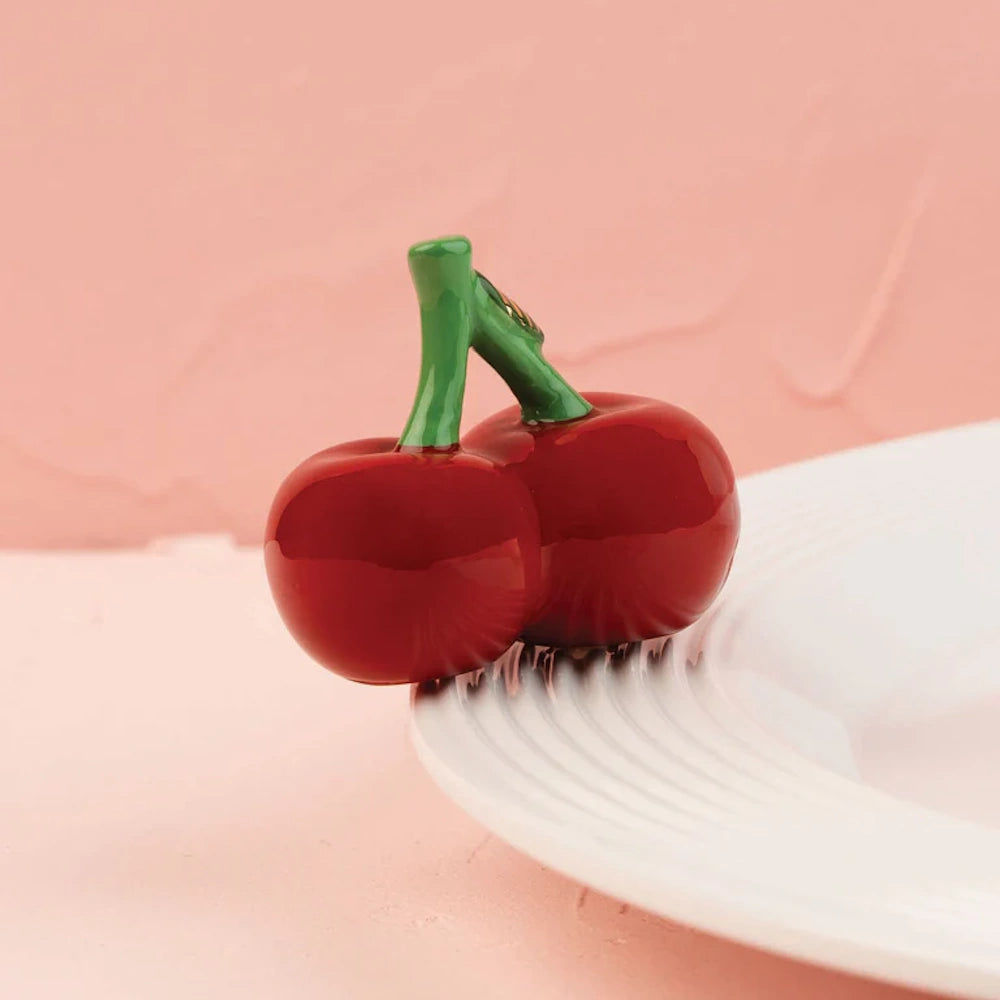 A glossy ceramic pair of red cherries with green stems resting on the rim of a white plate.