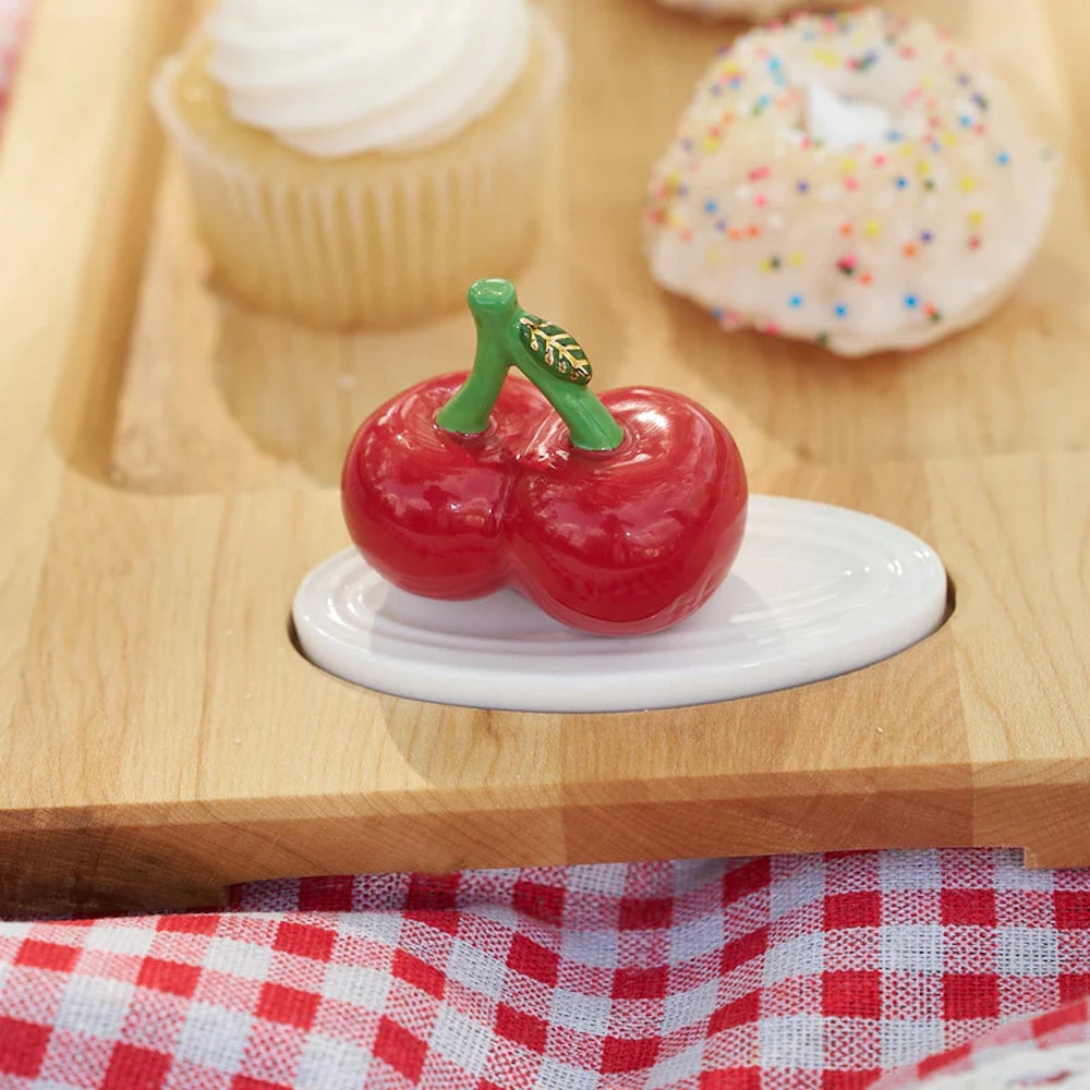 Small glossy ceramic double cherry charm with green stem set on a wooden serving tray beside treats.
