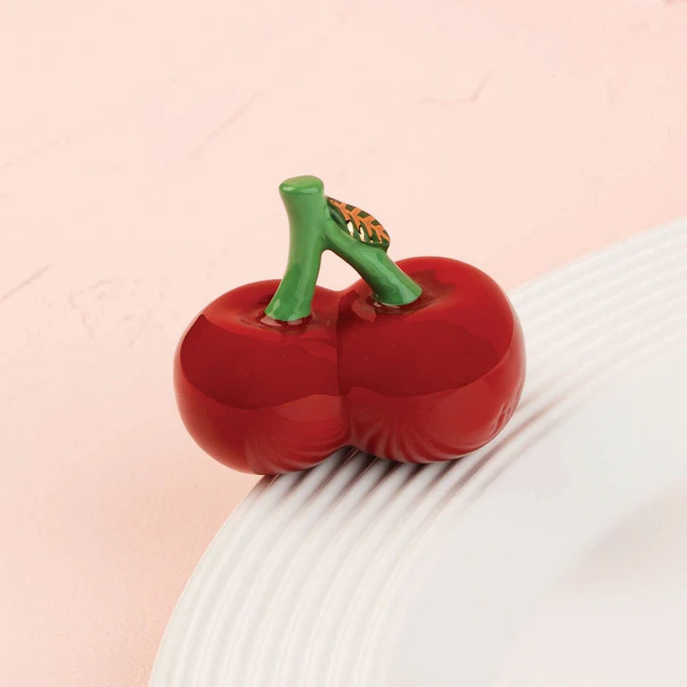 Glossy ceramic pair of ripe red cherries with green stems resting on the edge of a white dish.