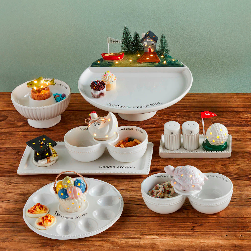 Collection of white ceramic serving dishes and light-up sitters with various themes on a wooden table.