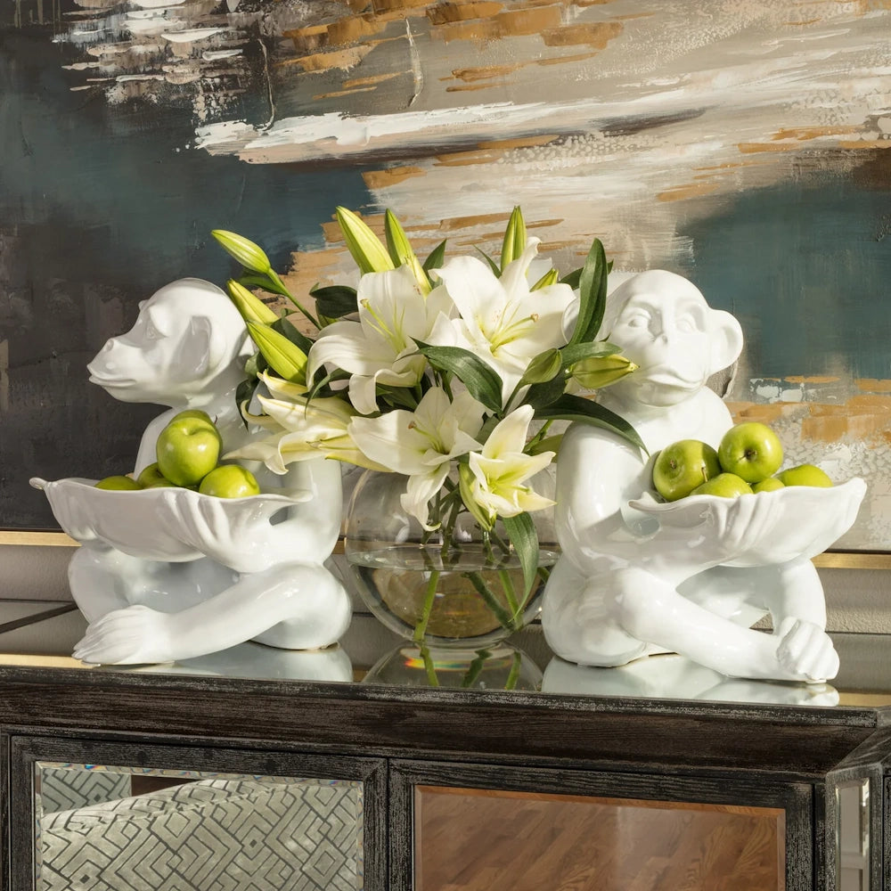 Two white ceramic monkey statues holding bowls of green apples next to a vase of white lilies.